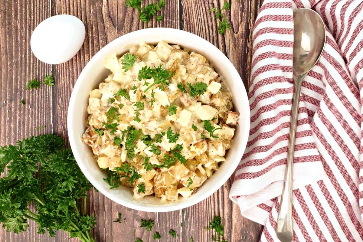A bowl of creamy potato salad garnished with chopped parsley sits on a wooden table next to a spoon, a striped towel, an egg, and fresh parsley.