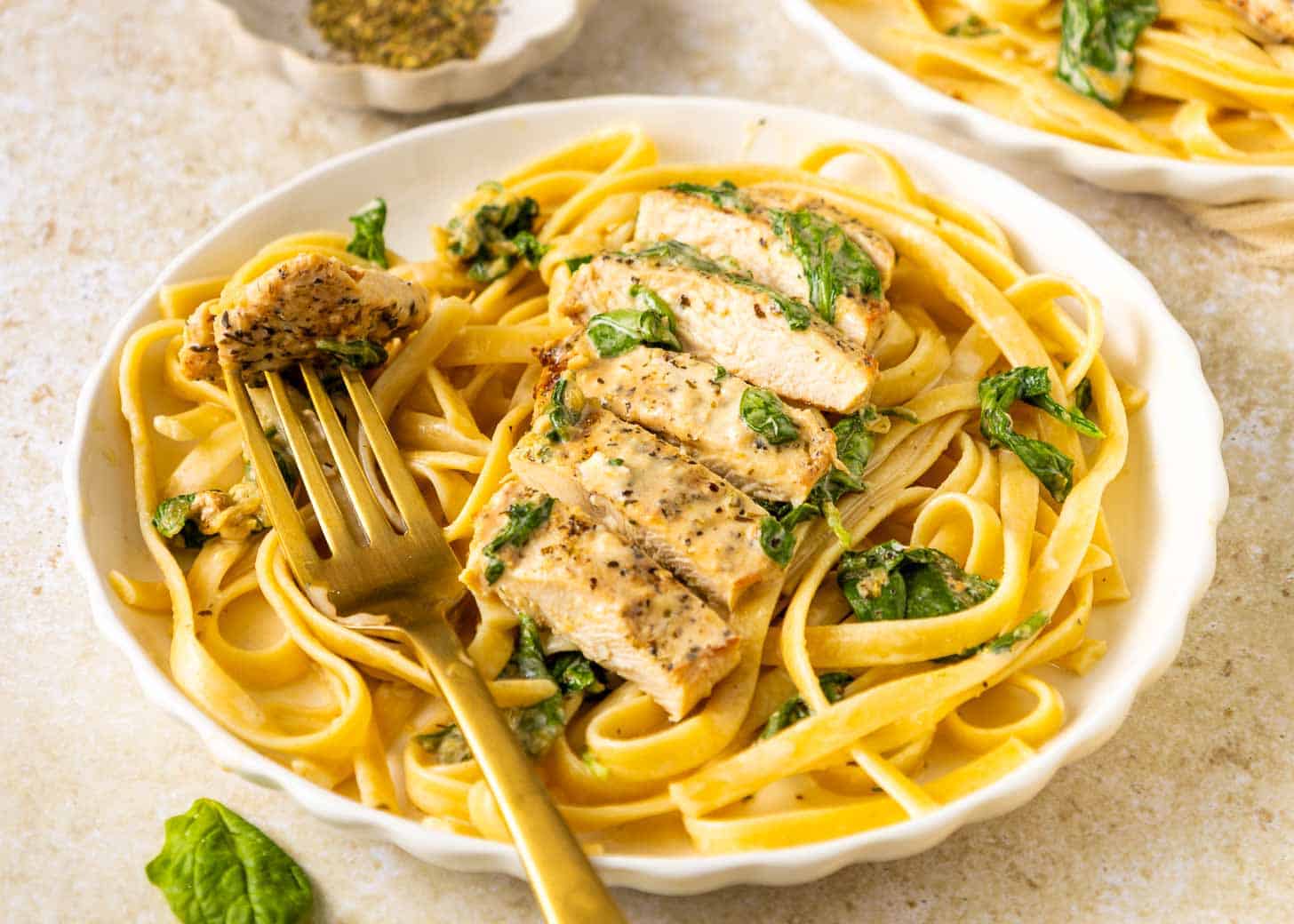 A plate of fettuccine pasta with spinach, topped with sliced grilled chicken breast, and a gold fork resting on the dish.