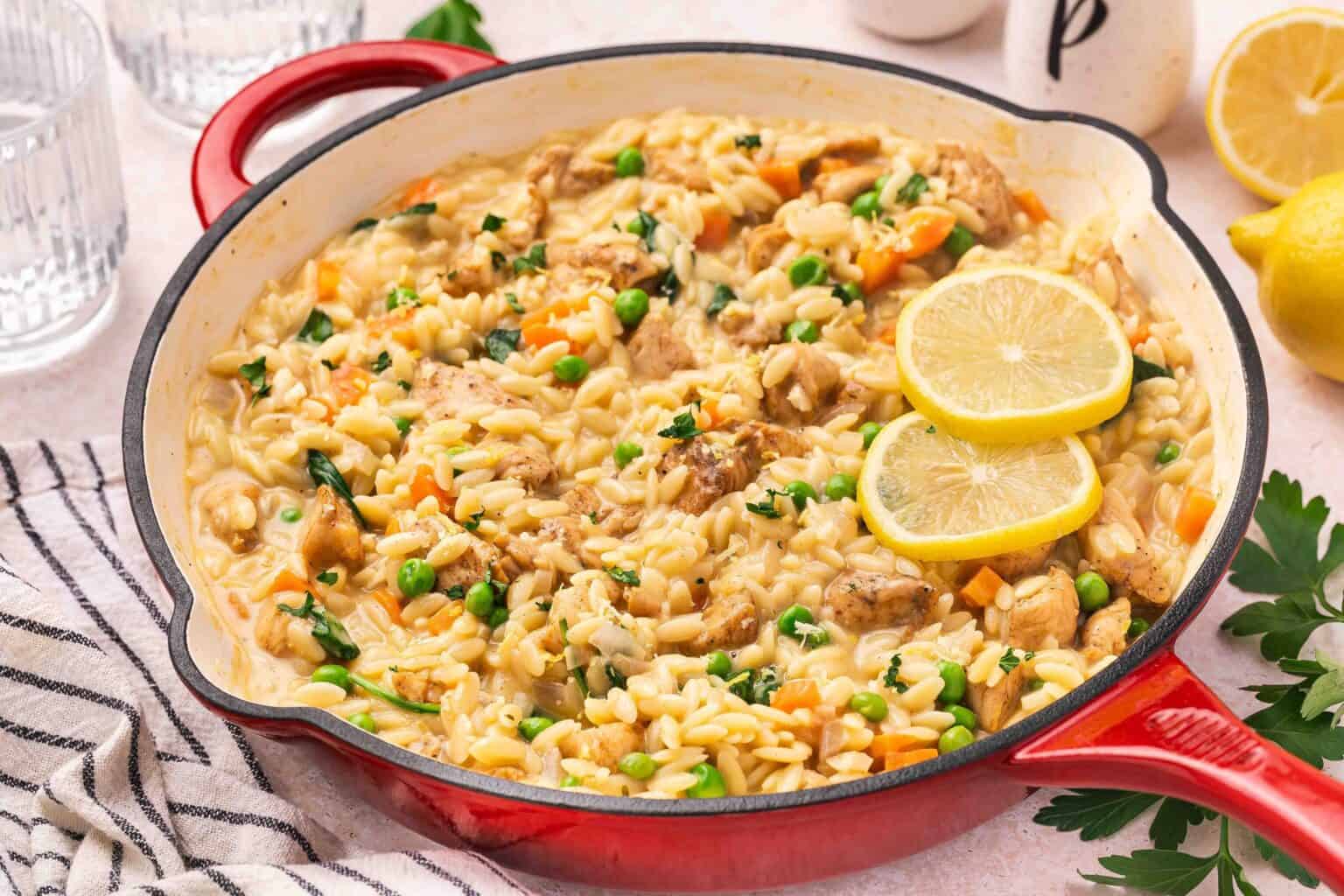A red skillet filled with creamy orzo pasta, peas, carrots, chicken pieces, and spinach, garnished with two lemon slices.