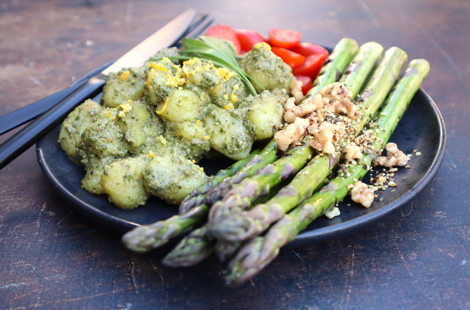 A plate with pesto gnocchi, fresh asparagus topped with chopped nuts, sliced cherry tomatoes, and a sprig of herbs, with a fork and knife beside the plate.