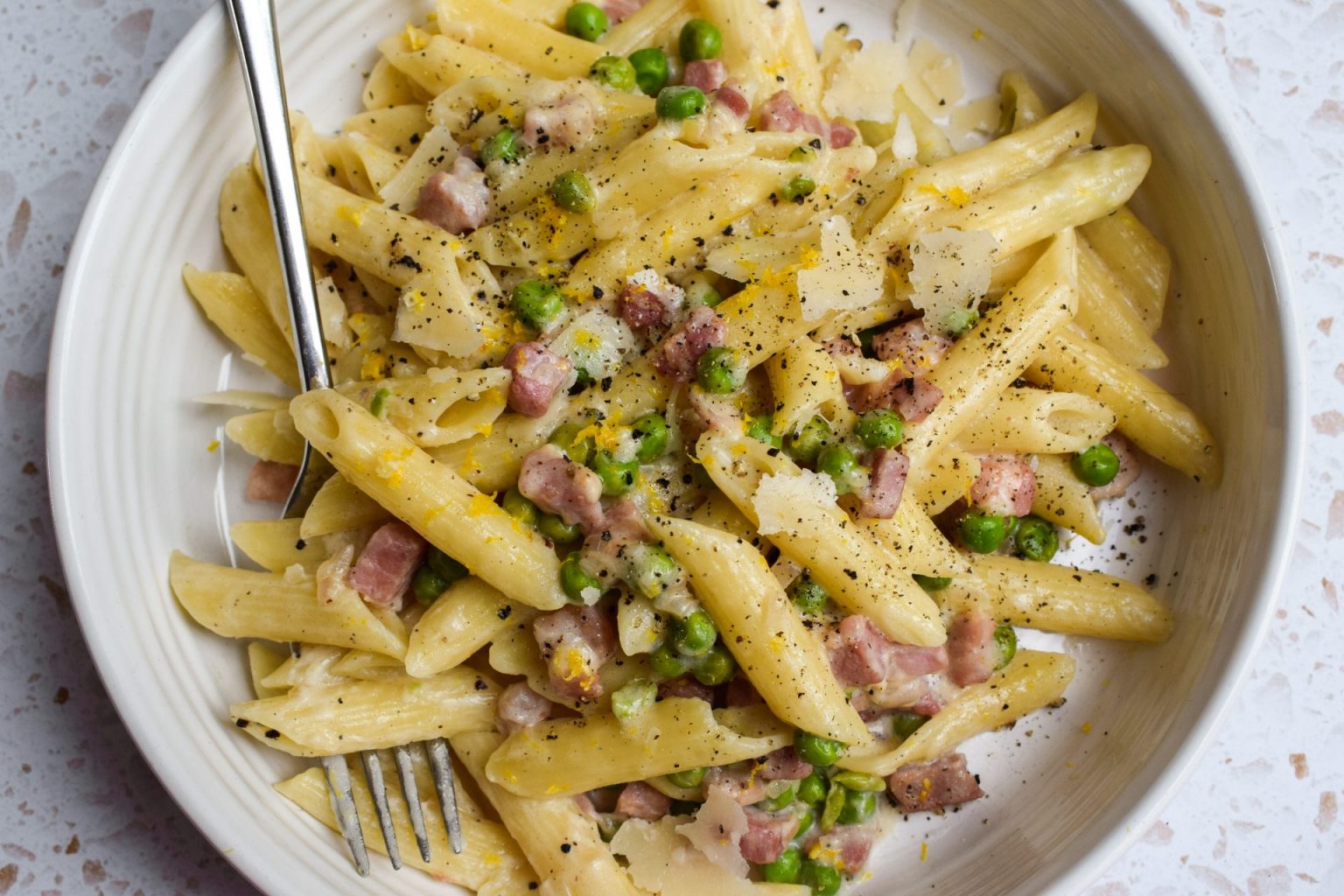 A bowl of penne pasta with diced ham, green peas, grated cheese, and black pepper, served with a fork.
