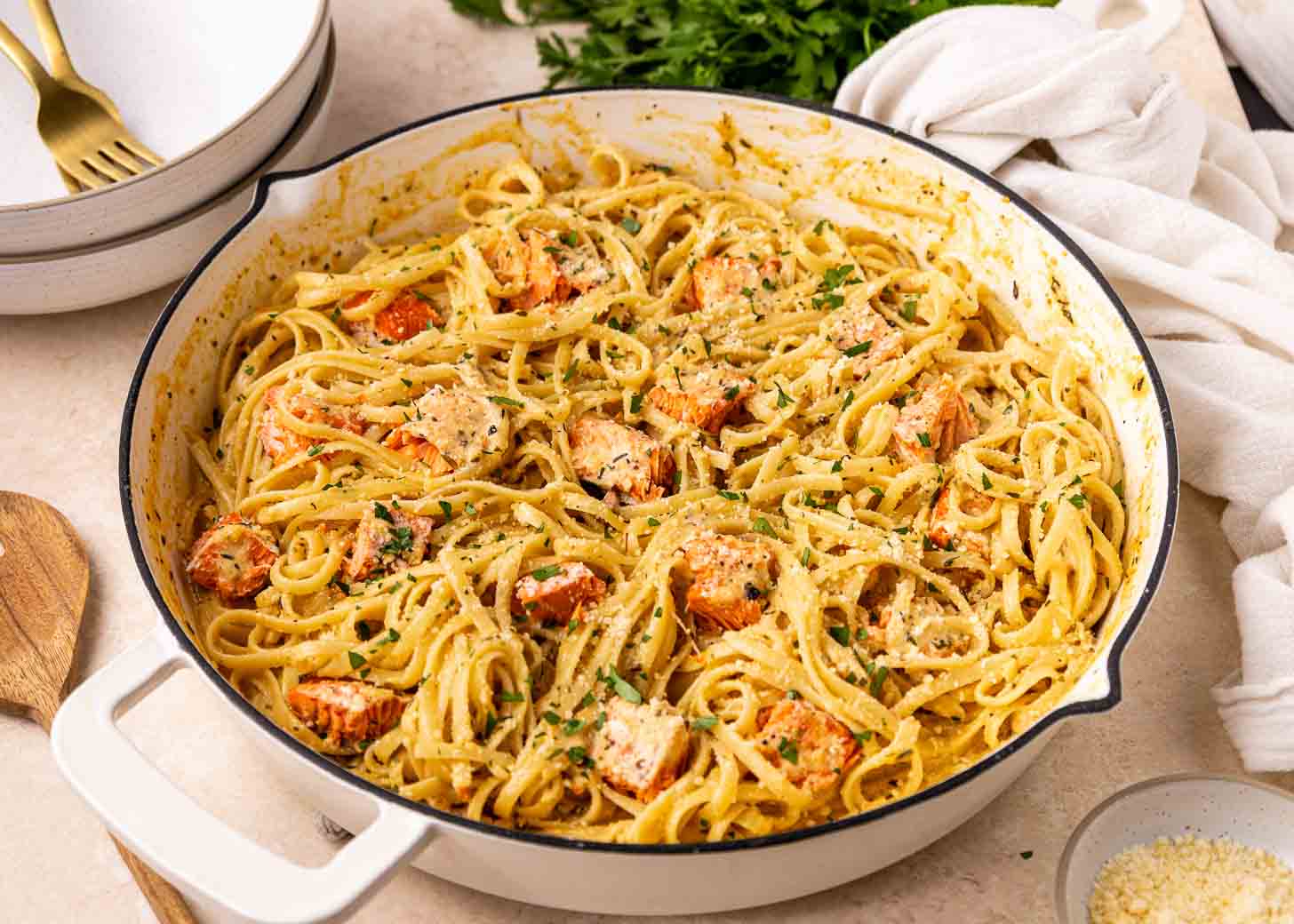 A large white skillet filled with creamy fettuccine pasta and chunks of salmon, garnished with chopped herbs and grated cheese.