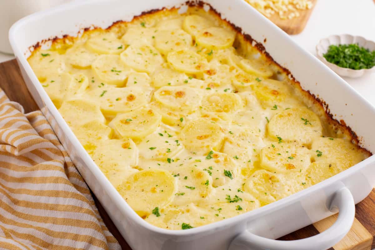 A white baking dish filled with baked scalloped potatoes topped with a light golden crust, with a striped towel and a bowl of chopped herbs nearby.