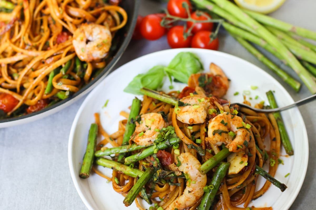 A plate of pasta with shrimp, asparagus, and cherry tomatoes garnished with herbs, next to a pan with more of the same dish and fresh vegetables.