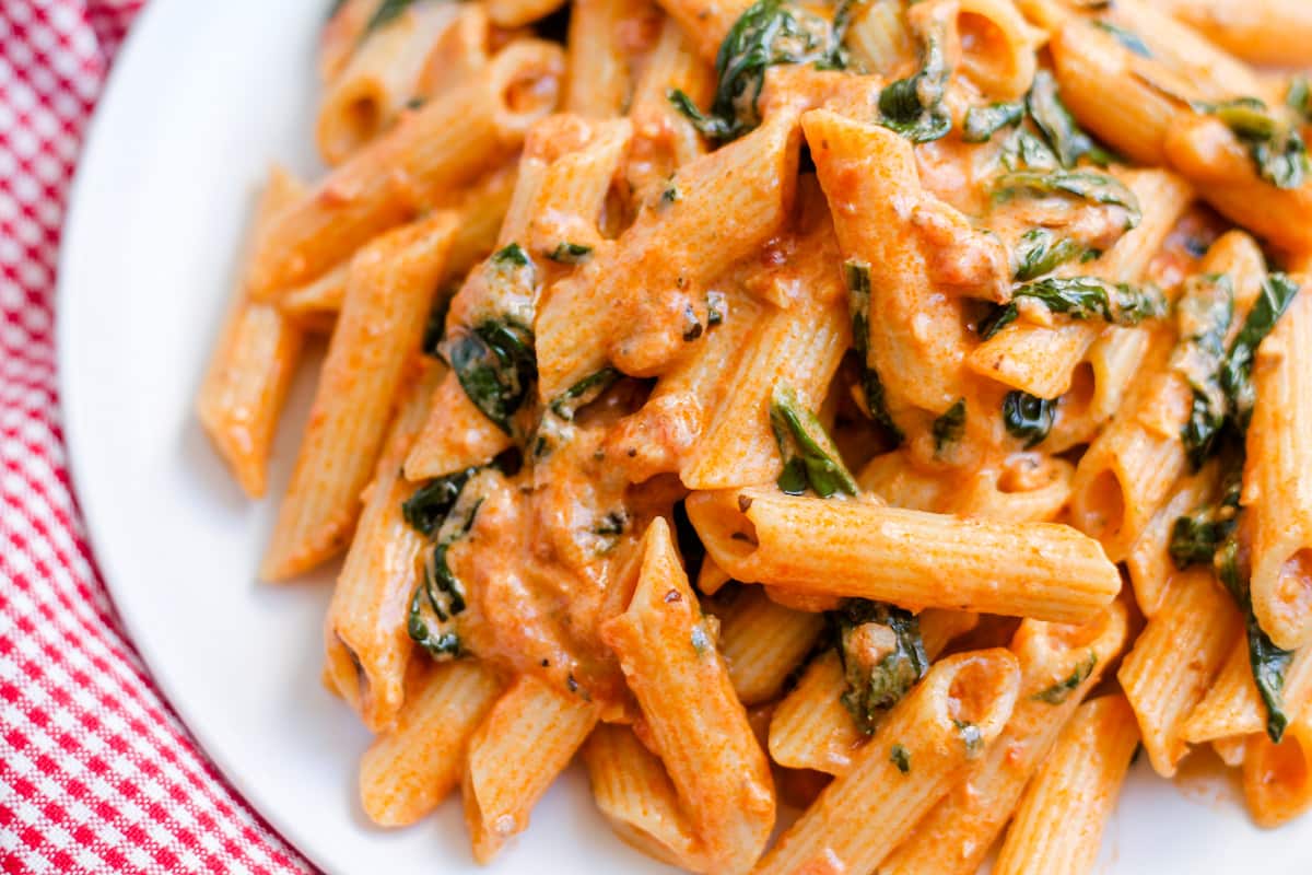 A plate of penne pasta with creamy tomato sauce and wilted spinach, served on a white dish over a red and white checkered cloth.