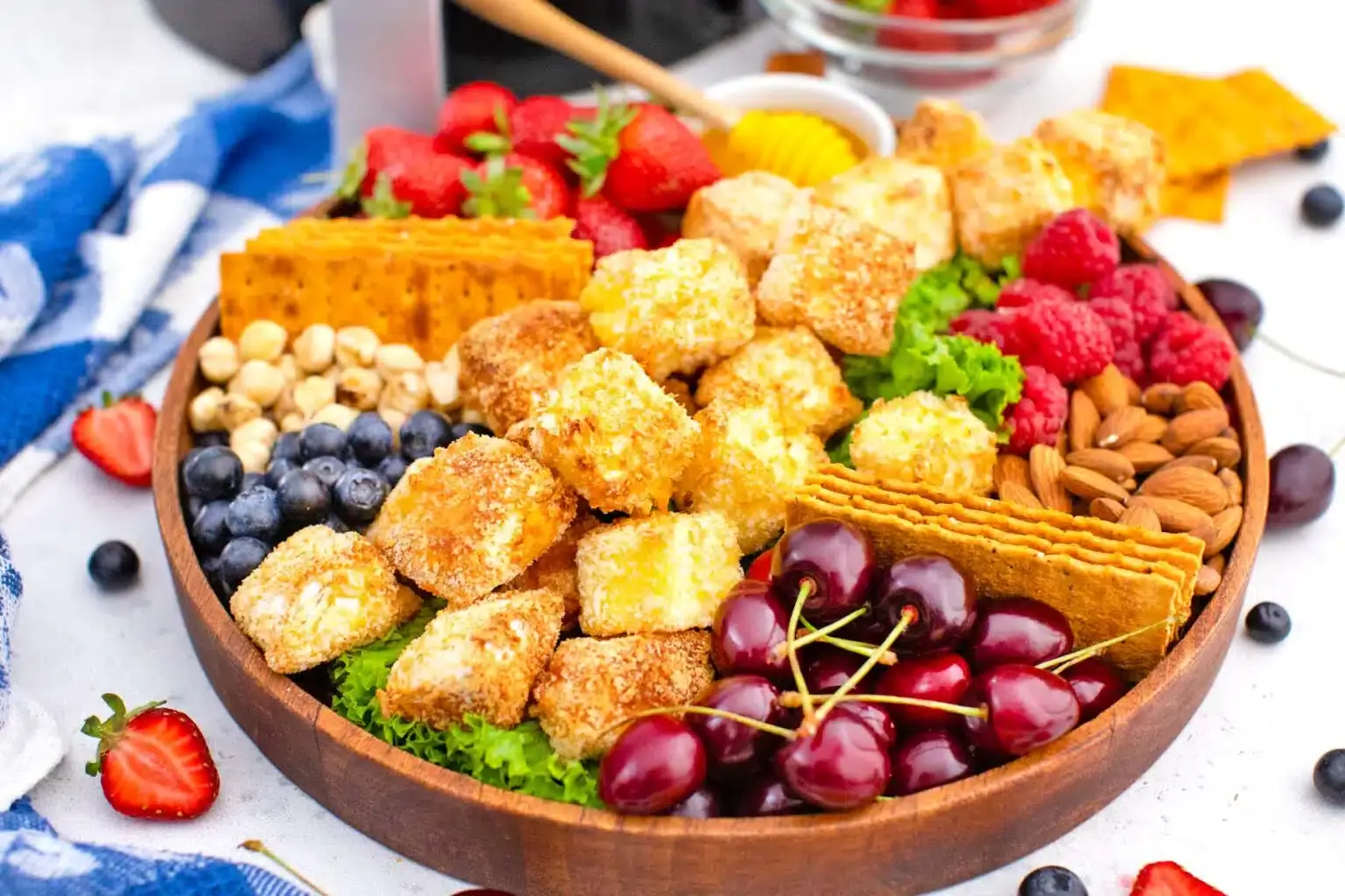 A wooden platter filled with berries, cherries, raspberries, nuts, crackers, and cubes of breaded cheese, arranged on a bed of leafy greens.