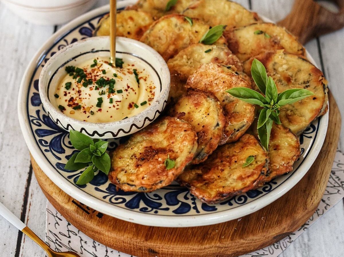 A plate of golden, fried zucchini slices garnished with fresh herbs, served with a bowl of creamy dip topped with chili flakes and chives on a patterned dish.