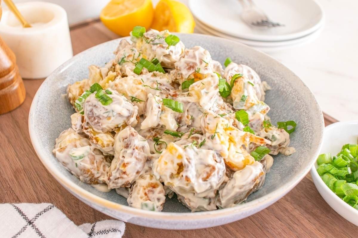 A bowl of creamy potato salad garnished with chopped green onions, placed on a wooden surface with lemon halves and plates in the background.