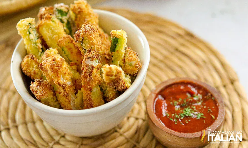 A white bowl filled with breaded zucchini fries sits on a woven placemat next to a small dish of marinara sauce.