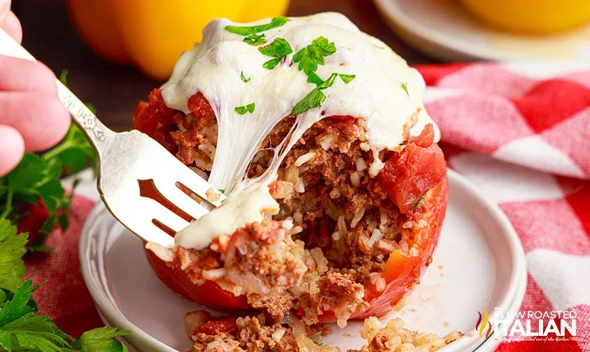 A fork cutting into a stuffed tomato filled with rice, ground meat, and topped with melted cheese, garnished with parsley, on a white plate.