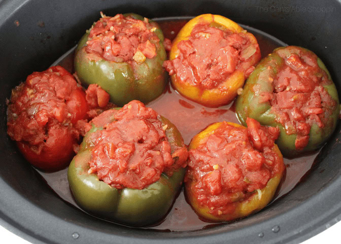 Six bell peppers, filled with a tomato-based mixture, are cooking in a black pot with tomato sauce.