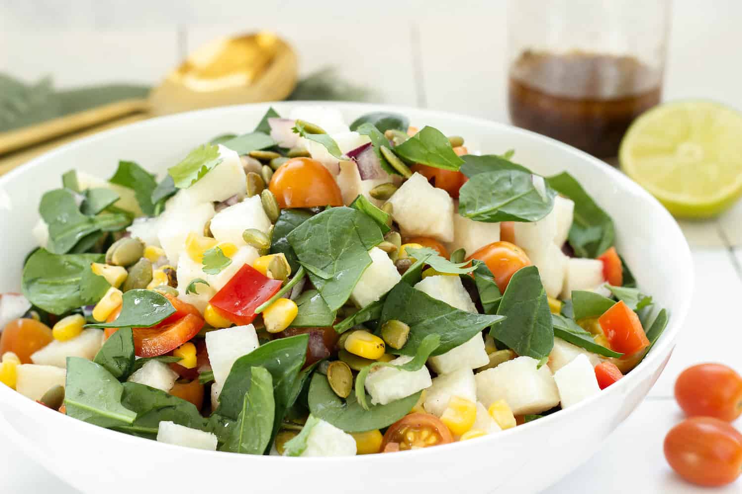A white bowl filled with a salad made of spinach, cherry tomatoes, jicama, red bell pepper, corn, and pumpkin seeds. A jar of dressing and a halved lime are in the background.