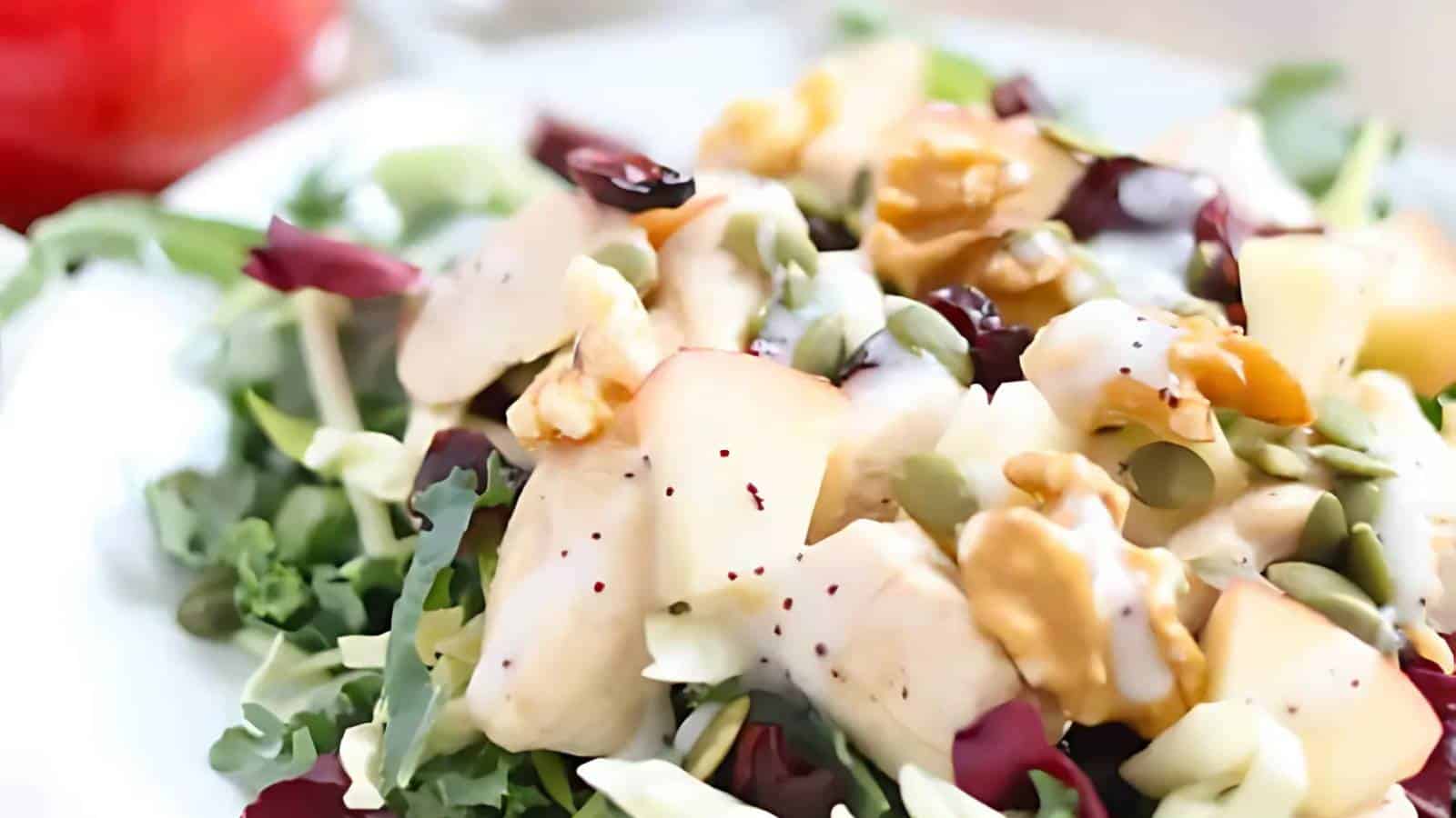 A close-up of a fresh salad with leafy greens, sliced apples, walnuts, seeds, and a light dressing on a white plate.
