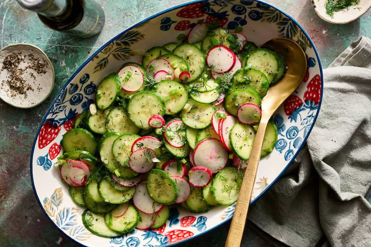 A large oval bowl filled with sliced cucumbers, radishes, and fresh herbs, served with a wooden spoon on a textured surface.