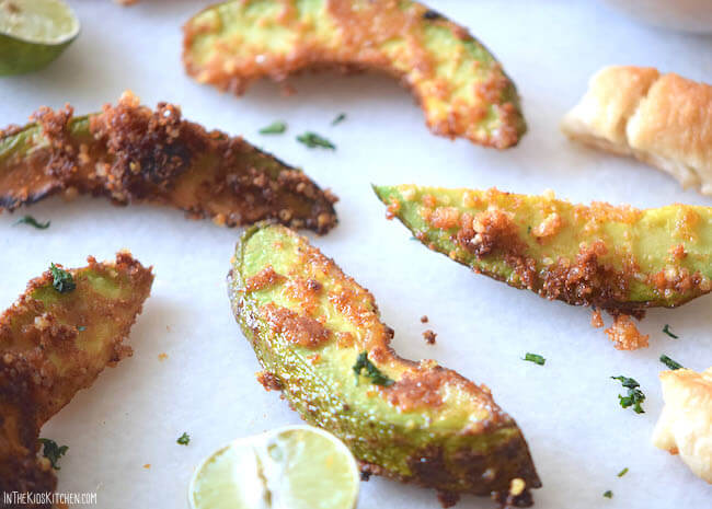 Sliced avocado wedges coated in breadcrumbs and spices, baked until golden brown, served on a white surface with lime wedges and herbs.