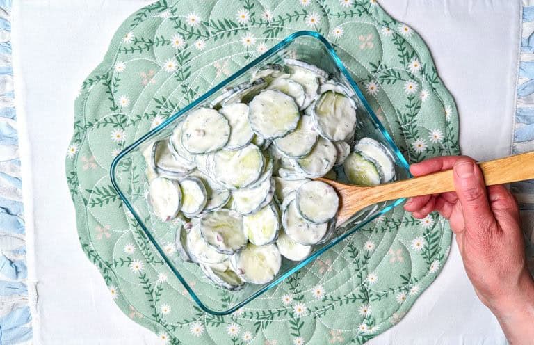 A hand holding a wooden spoon over a glass dish filled with creamy cucumber salad, placed on a green patterned placemat.