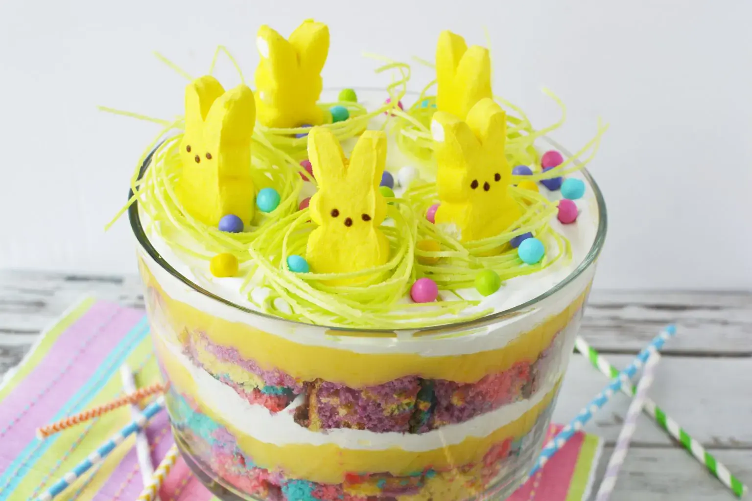 A layered trifle dessert topped with yellow marshmallow bunny candies, green edible grass, and colorful round candies, set on a pastel napkin with striped straws beside it.