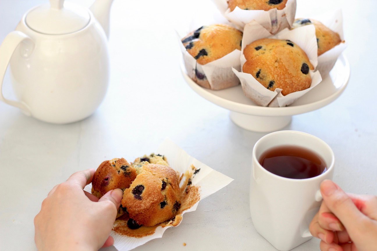 A person holds a blueberry muffin on a napkin while another hand holds a cup of tea; a teapot and more muffins are on a white cake stand in the background.