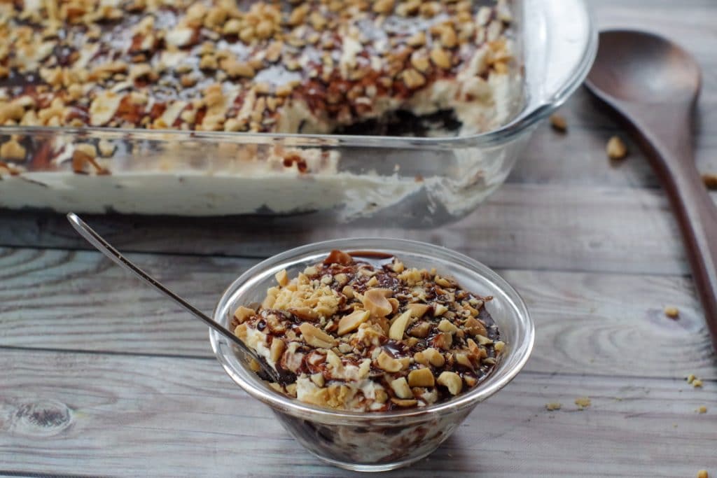 A glass dish and a small bowl filled with layered dessert topped with chopped nuts and chocolate sauce, with a spoon in the bowl.