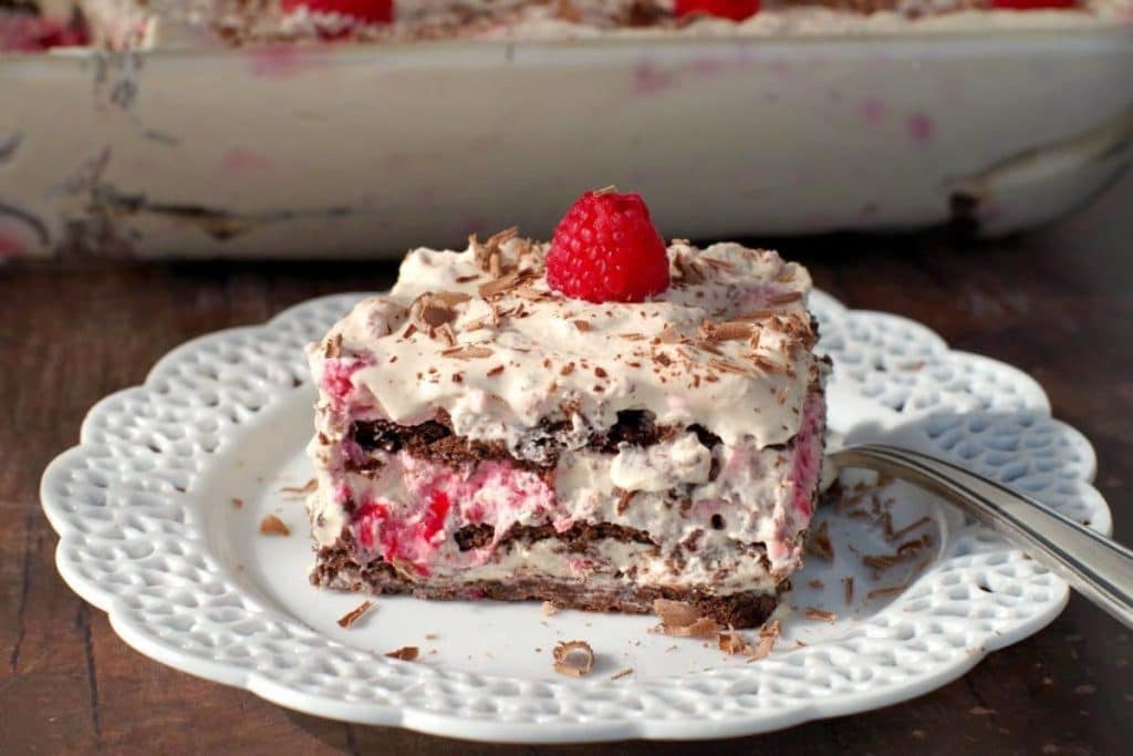 A slice of layered dessert with whipped cream, chocolate, and raspberries sits on a white plate with a fork, topped with a single raspberry and chocolate shavings.