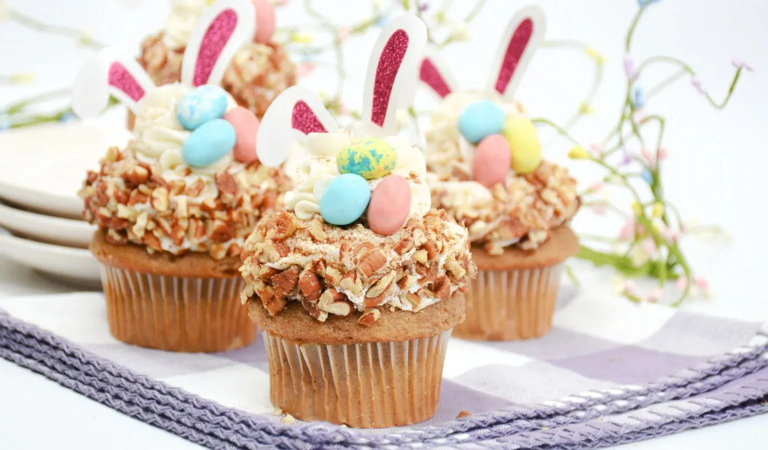 Three cupcakes topped with chopped nuts, pastel candy eggs, and bunny ear decorations sit on a checkered napkin with plates in the background.