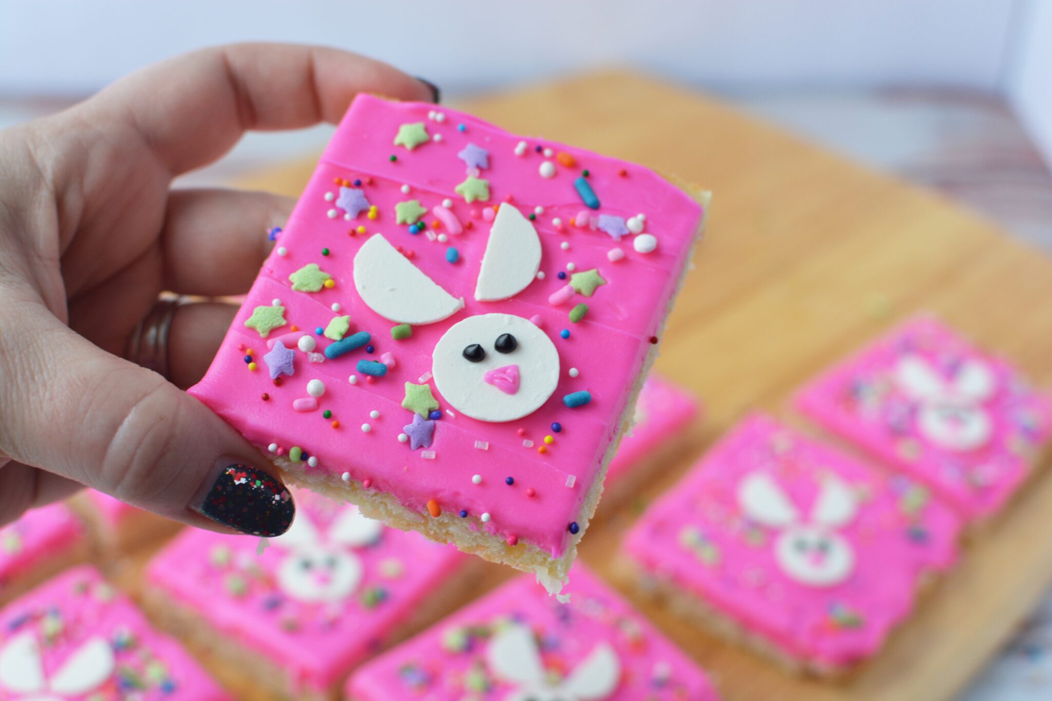 A hand holds a pink frosted cookie bar decorated with a bunny face and colorful sprinkles; similar bars are on a wooden surface in the background.