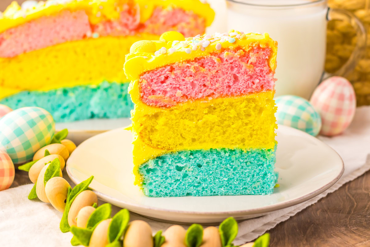 A slice of three-layer cake in pink, yellow, and blue with yellow frosting on a white plate, surrounded by Easter eggs and a glass of milk.