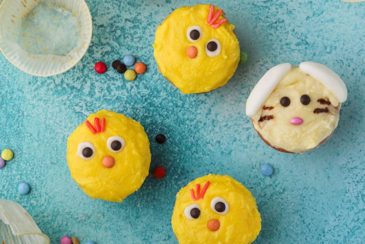 Three cupcakes decorated as yellow chicks and one cupcake decorated as a white bunny are arranged on a blue surface, with scattered colorful candies around them.