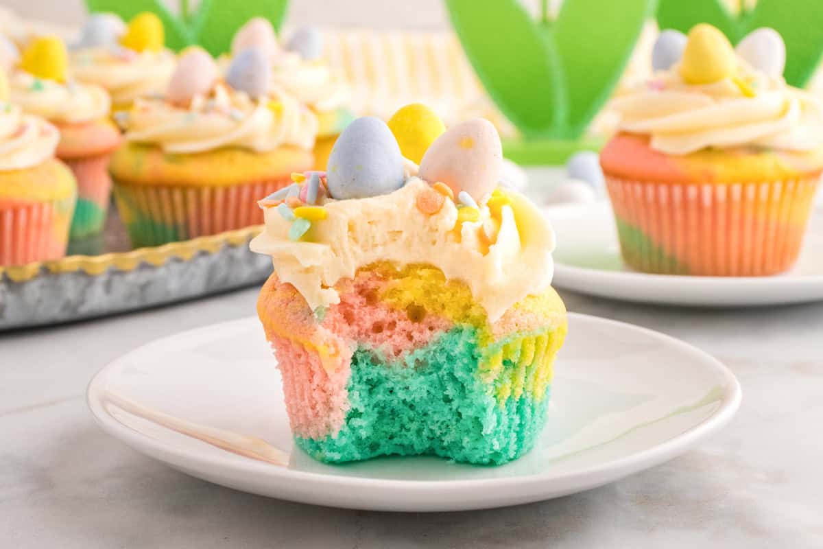 A pastel-colored cupcake with a bite taken out, topped with white frosting, sprinkles, and candy eggs, sits on a white plate. More decorated cupcakes are in the background.
