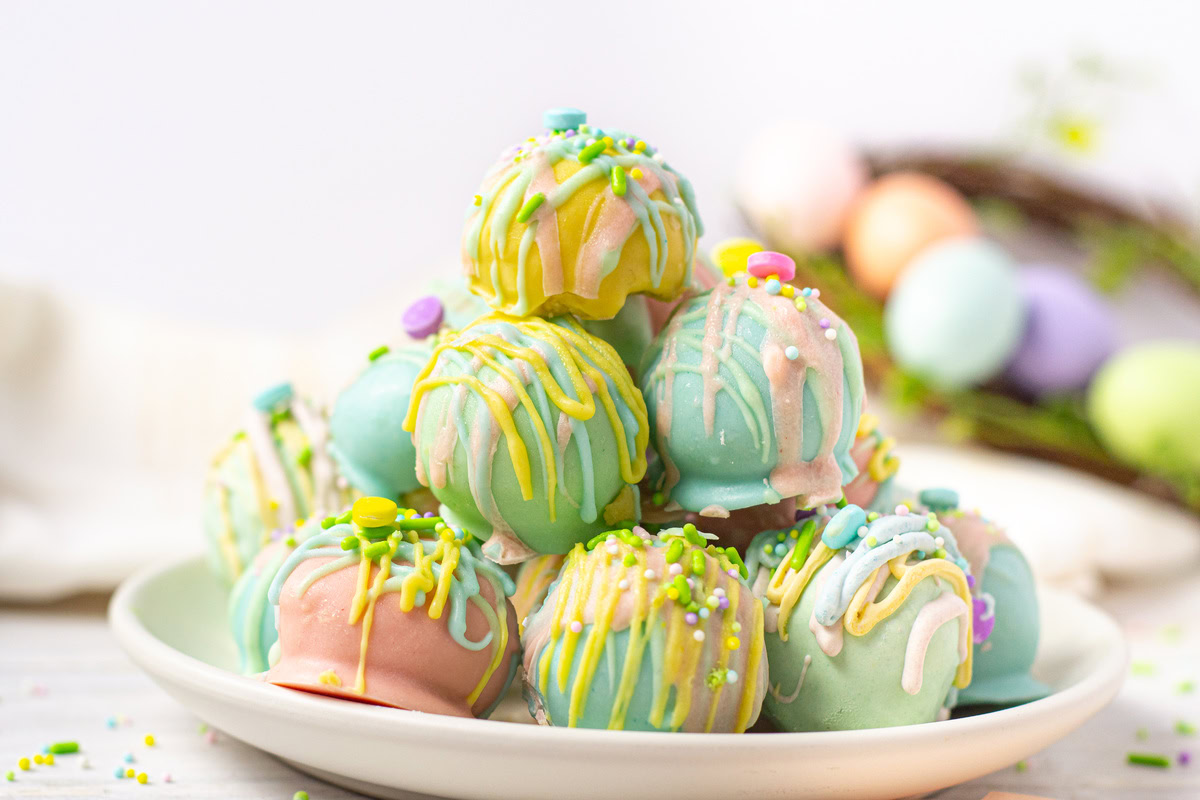 A plate of pastel-colored cake balls decorated with colorful drizzle, sprinkles, and small candy pieces, with blurred Easter eggs in the background.