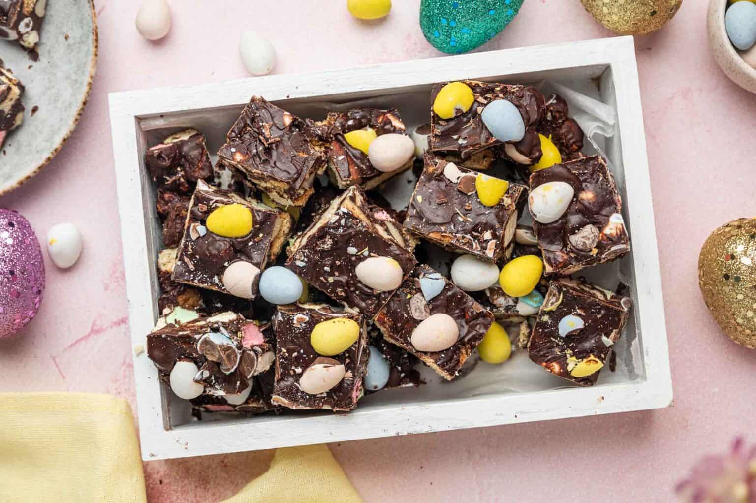 A white tray filled with chocolate bark pieces topped with colorful candy-coated eggs, surrounded by decorative eggs on a pink surface.