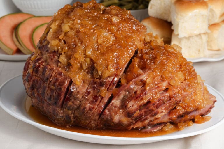 A cooked ham topped with a chunky pineapple glaze is served on a white platter, with bread rolls and melon slices in the background.