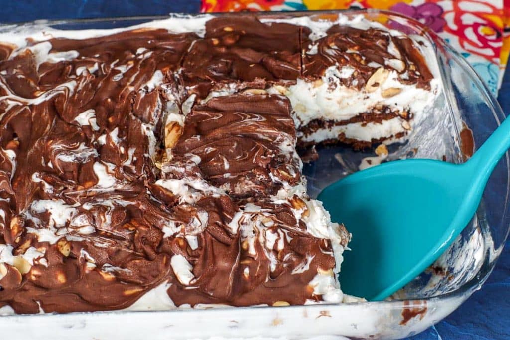 A glass dish filled with layered chocolate and cream dessert, partially served with a blue spatula, sits on a colorful cloth.
