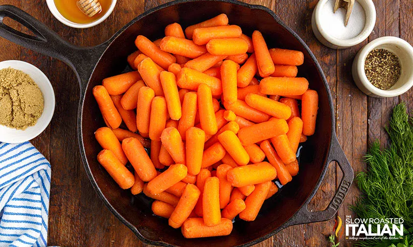 A cast iron skillet filled with baby carrots sits on a wooden table surrounded by bowls of brown sugar, salt, pepper, honey, fresh dill, and a striped towel.