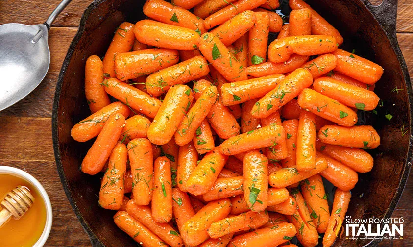 A skillet filled with glazed baby carrots garnished with chopped herbs, next to a bowl of honey and a spoon on a wooden surface.