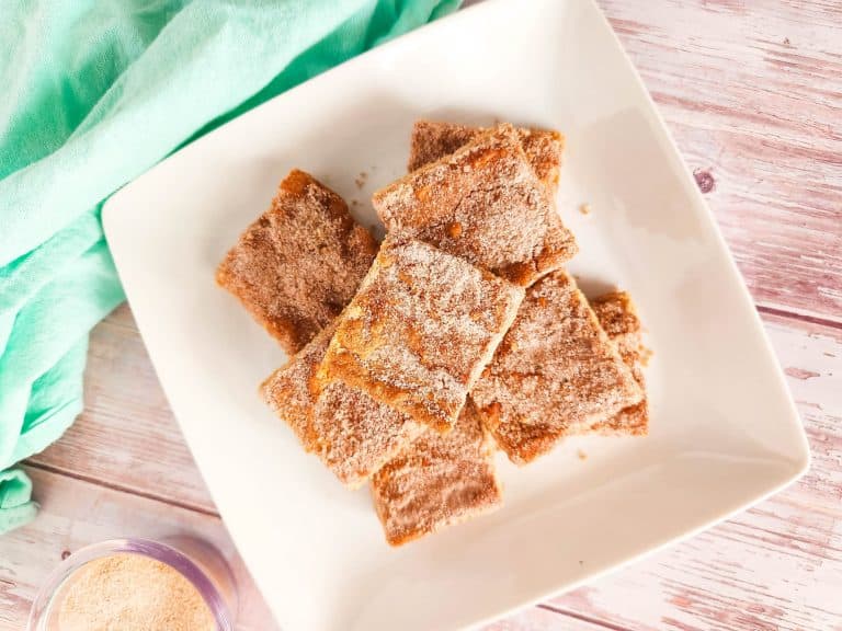 A white plate with a stack of cinnamon-sugar coated bars sits on a light wooden surface next to a light green cloth.