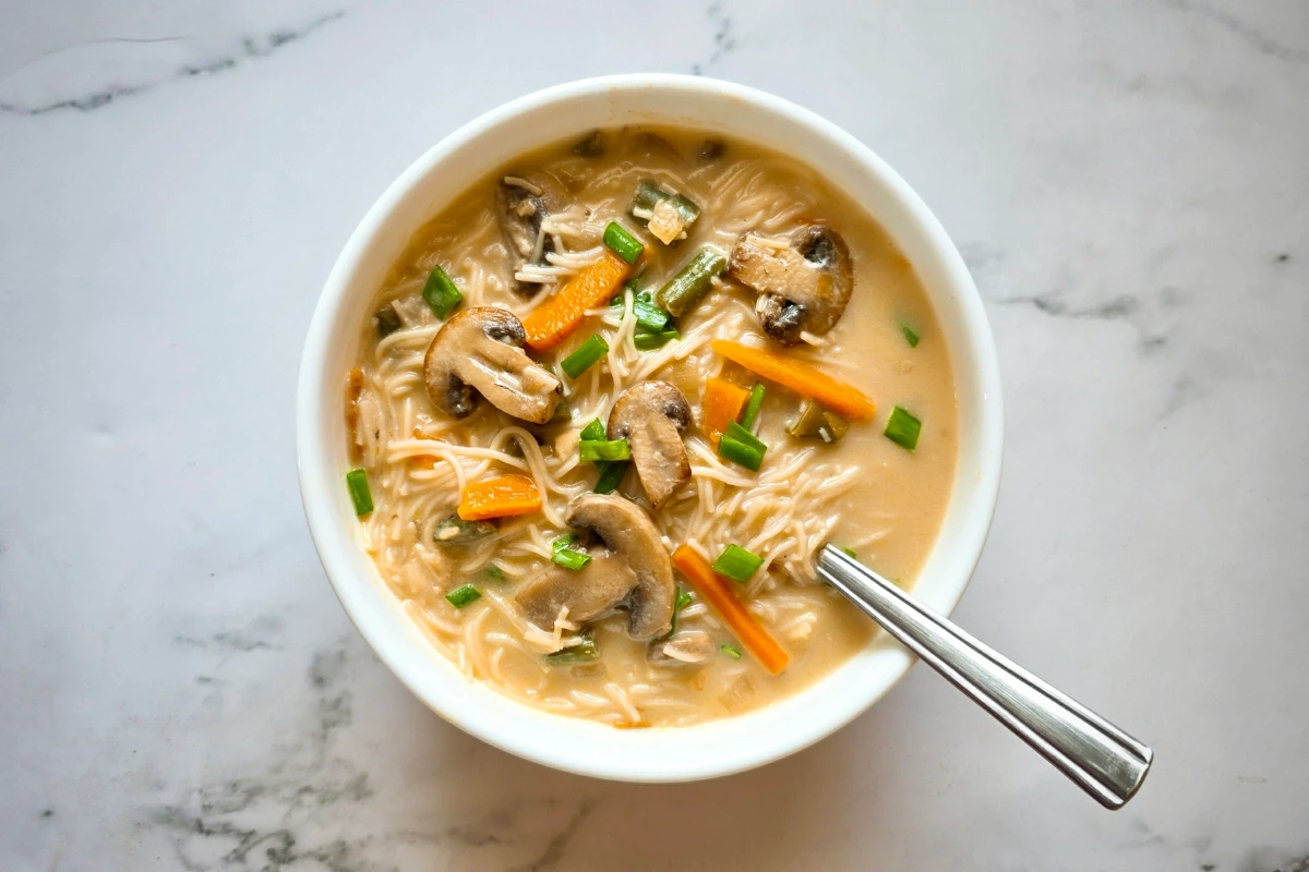 A bowl of noodle soup with mushrooms, carrots, green onions, and a spoon, on a white marble surface.