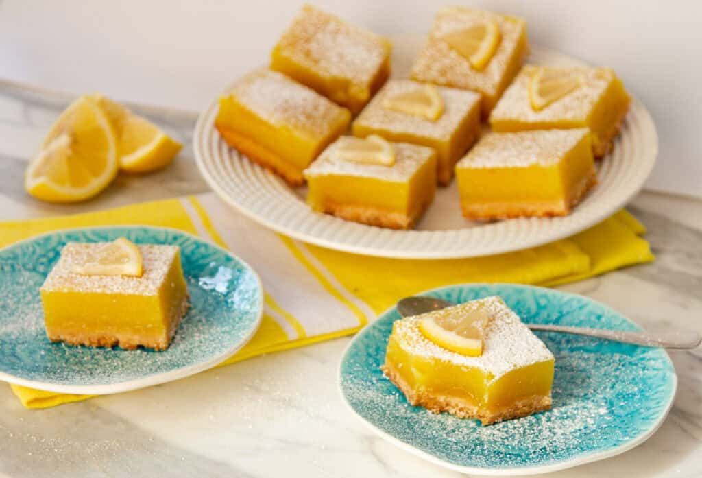 Lemon bars topped with powdered sugar and lemon slices are served on blue plates and a white platter, with lemon wedges in the background.