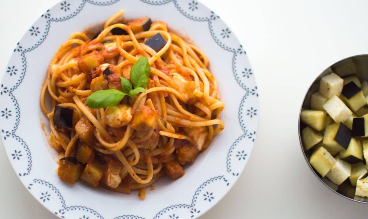 A plate of spaghetti with tomato sauce, diced eggplant, and basil garnish, next to a bowl of raw cubed eggplant on a white surface.