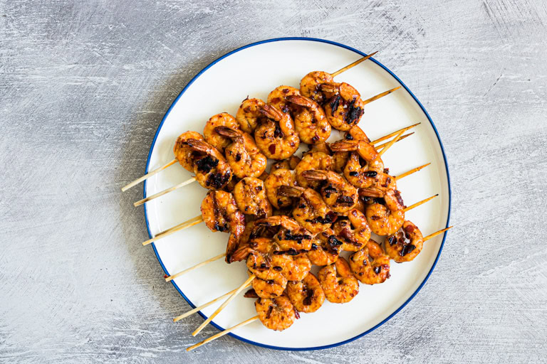A plate of grilled shrimp skewers arranged in a fan shape on a white plate with a blue rim, set on a light textured surface.