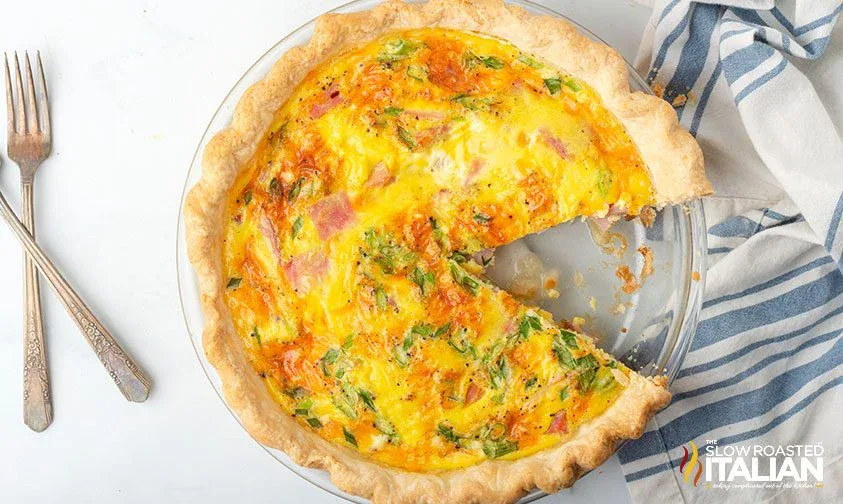 A baked quiche with a slice missing, filled with vegetables and ham, sits in a glass pie dish next to a striped napkin and two forks.
