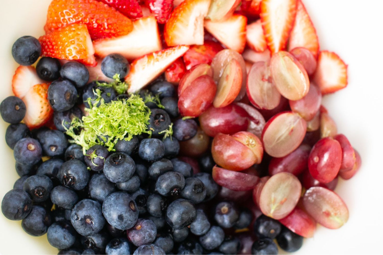 A close-up of fresh blueberries, halved strawberries, sliced red grapes, and a small pile of lime zest in a white bowl.