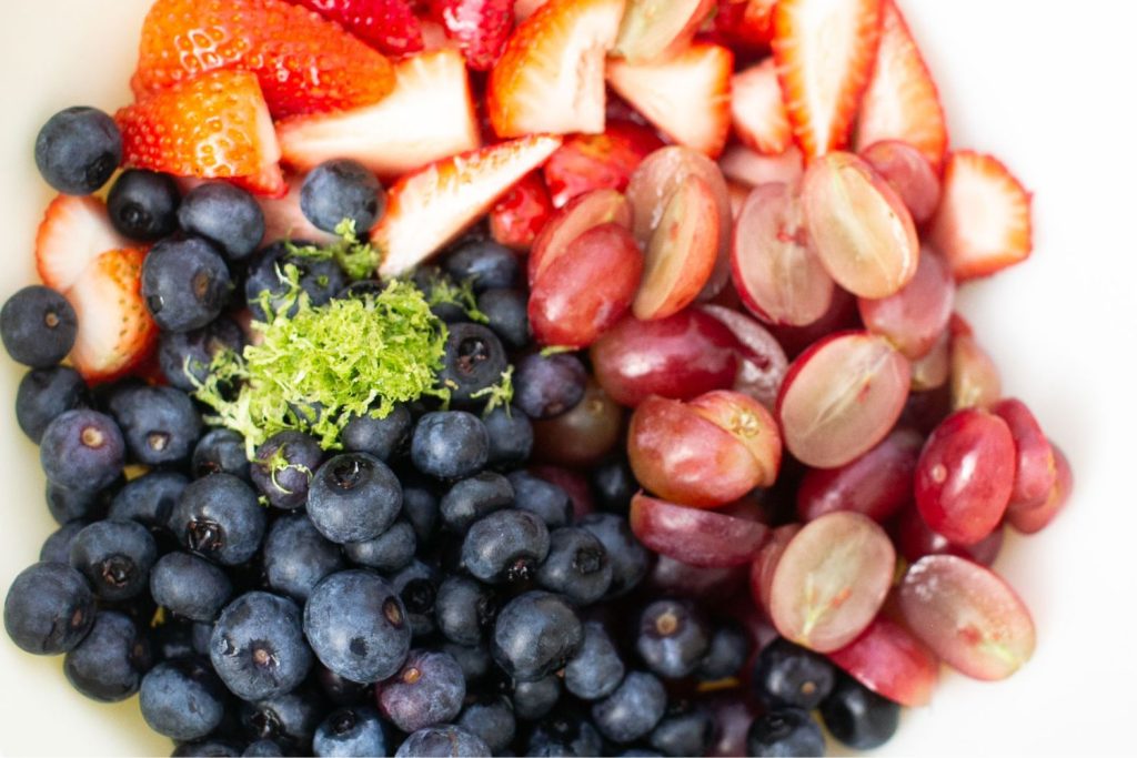 A close-up of fresh blueberries, halved red grapes, sliced strawberries, and a small pile of lime zest in a white bowl.