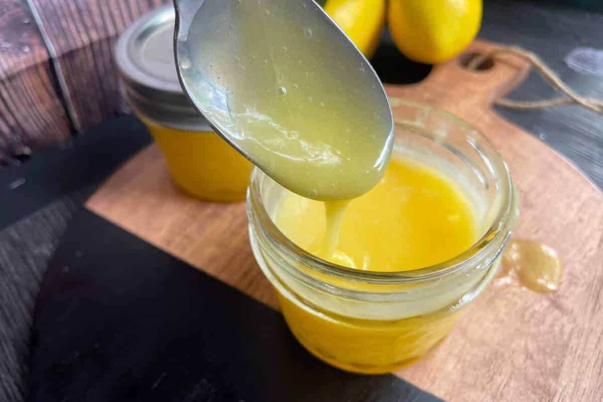 A spoon holds a thick, yellow lemon curd over an open glass jar on a wooden board. Another sealed jar and whole lemons are visible in the background.