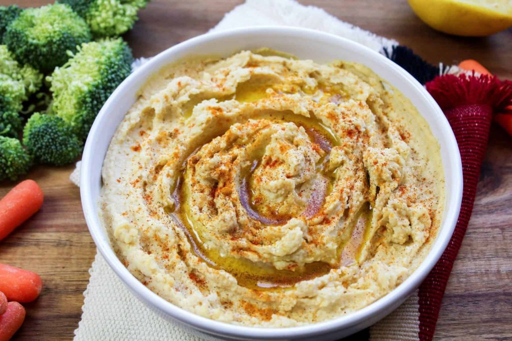 A bowl of hummus drizzled with olive oil and sprinkled with paprika, surrounded by broccoli florets and baby carrots on a wooden surface.