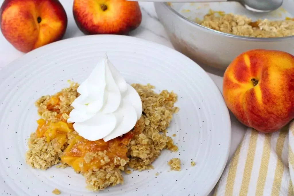 A serving of peach crumble topped with whipped cream on a white plate, surrounded by fresh peaches and a bowl of crumble in the background.