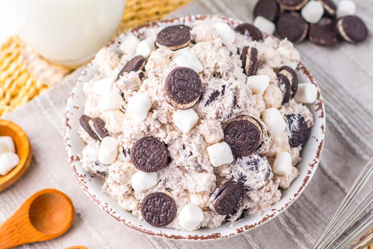 A bowl of Oreo fluff dessert topped with mini Oreo cookies and marshmallows, placed on a table with wooden measuring spoons and a glass of milk nearby.