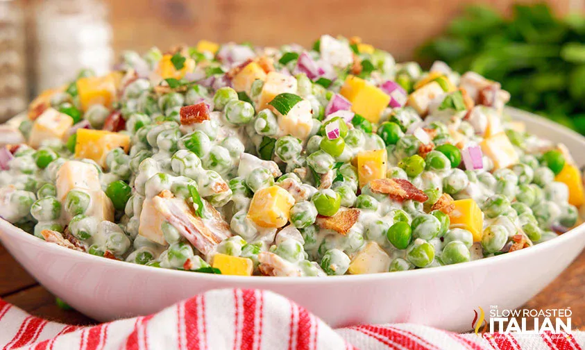 A bowl of creamy pea salad with diced cheddar cheese, bacon pieces, chopped red onion, and parsley.