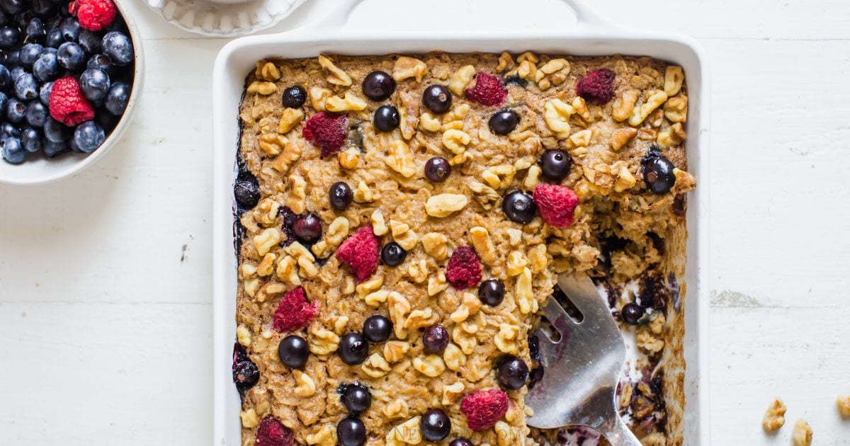 A baked oatmeal dish topped with walnuts, blueberries, and raspberries, with one serving missing and a metal spatula in the dish. A bowl of mixed berries sits nearby.