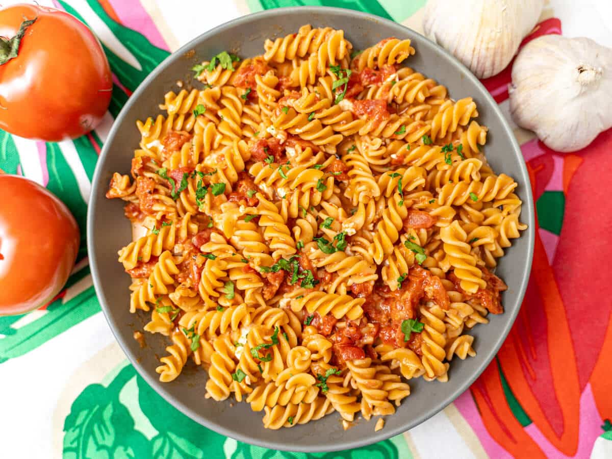 A plate of rotini pasta with tomato sauce, garnished with chopped parsley, sits on a colorful cloth next to two tomatoes and two heads of garlic.